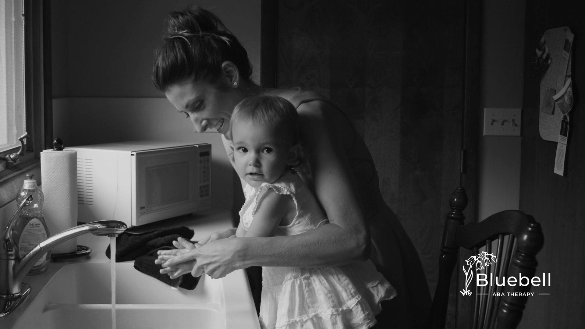 A BCBA helps a baby with autism wash hands at a kitchen sink during home-based ABA therapy in NC.