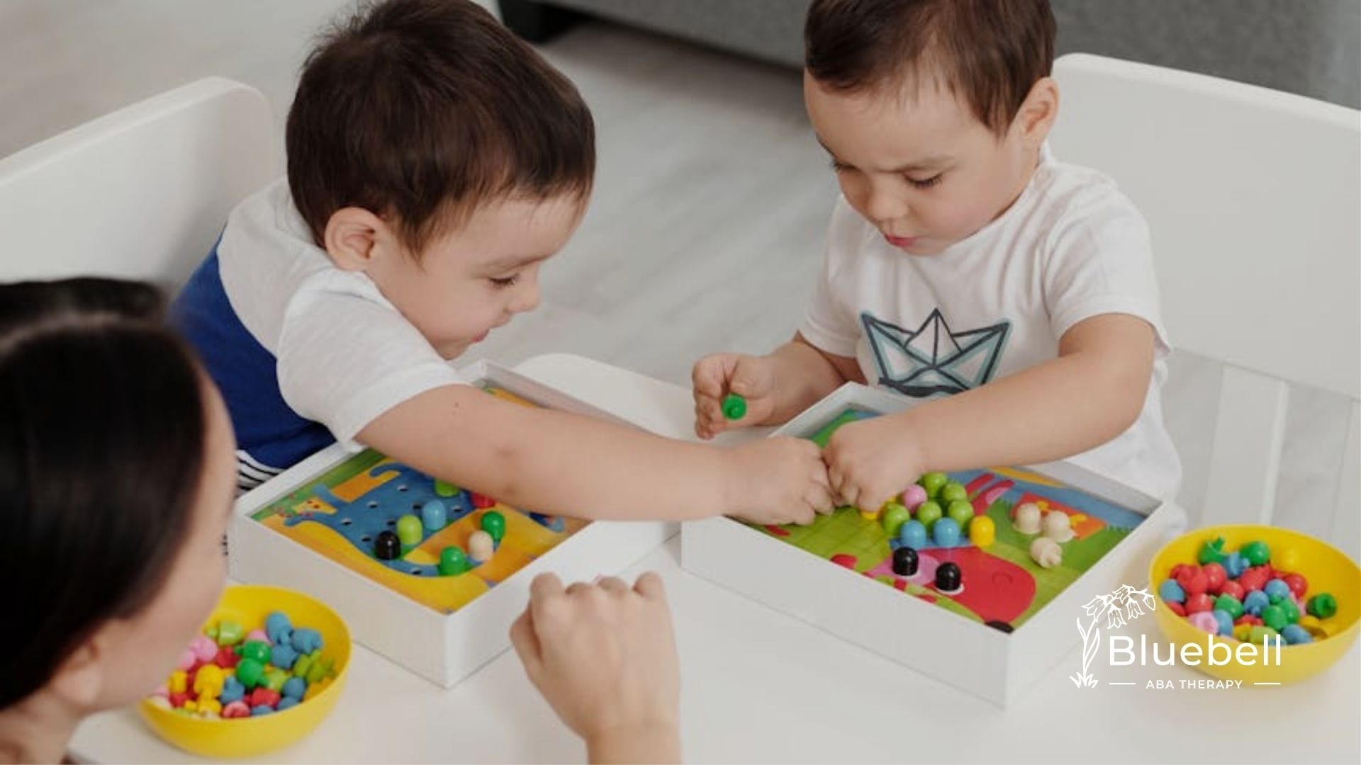 Two autistic toddlers play with colorful toys at a table while an ABA therapist assists them in NC.