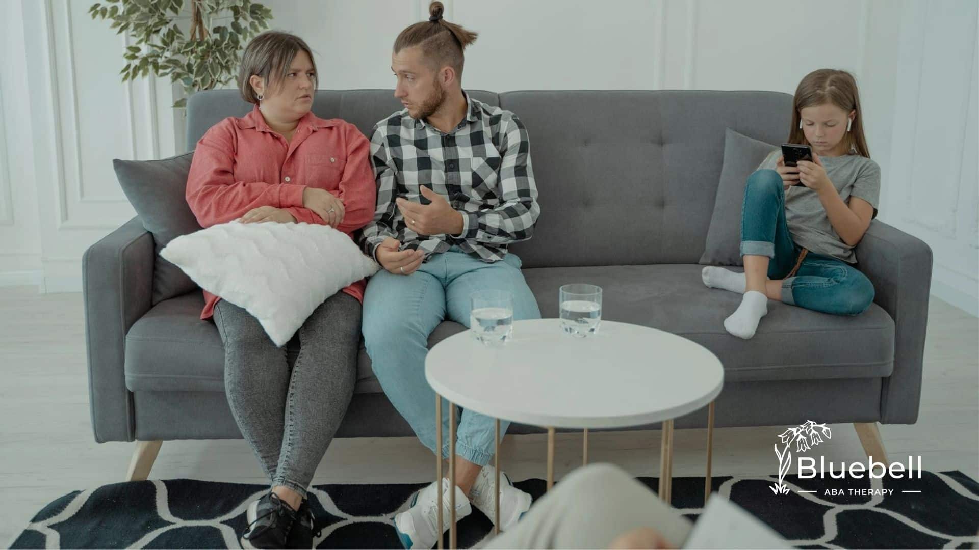 Parents discussing what questions to ask to ABA therapist while autistic daughter plays on a phone.