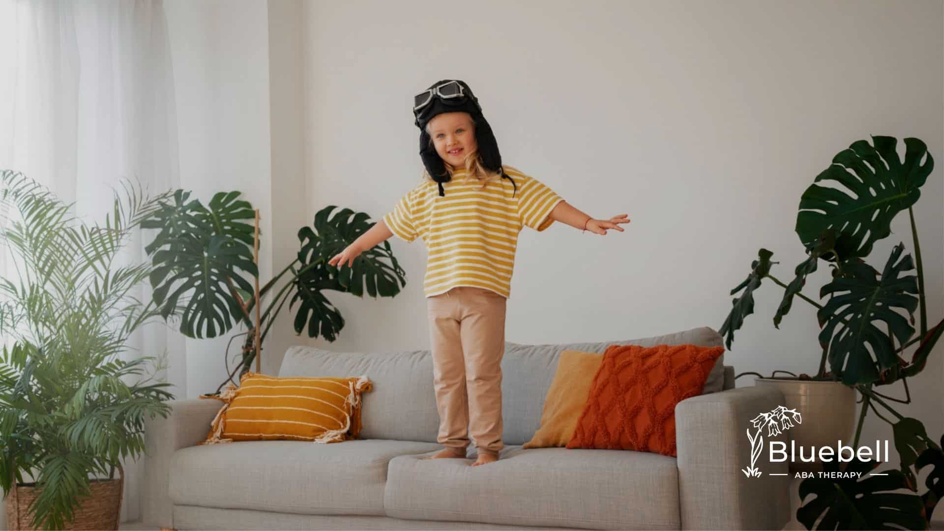 Autistic girl wearing a striped yellow shirt and aviator hat balances on a couch.