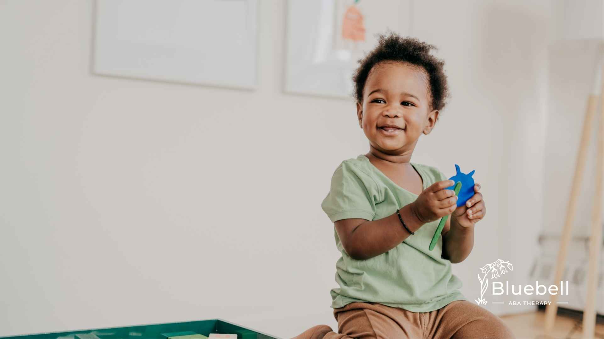 A joyful toddler in a green shirt sits indoors, holding a blue toy. The background is softly blurred, conveying a warm and playful atmosphere.