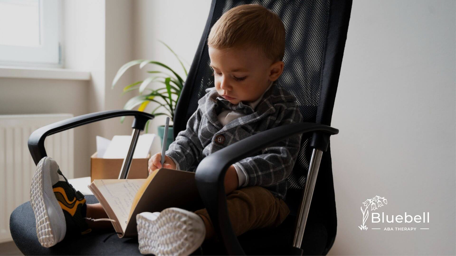 A toddler with autism dressed in a checked suit writes in a notebook while seated in an office chair.