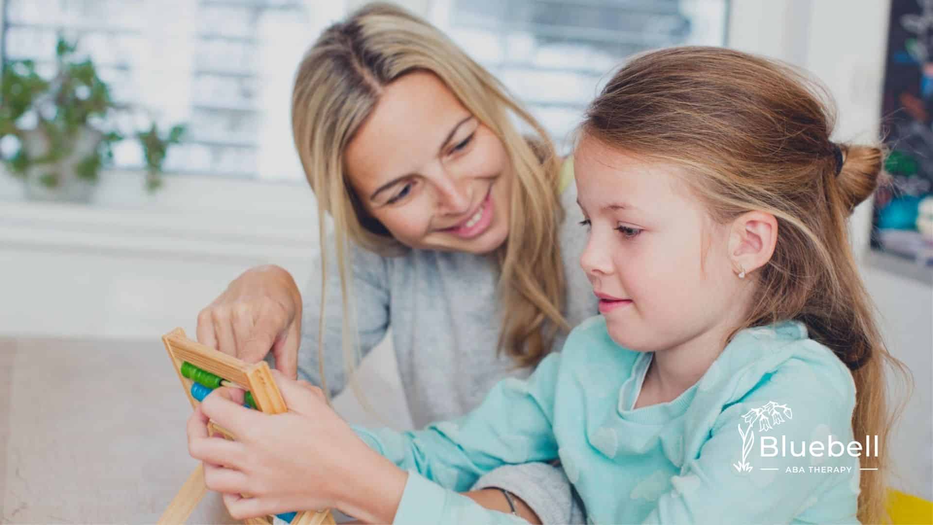 A BCBA helps a young girl with autism use a small wooden abacus at a table during ABA therapy.